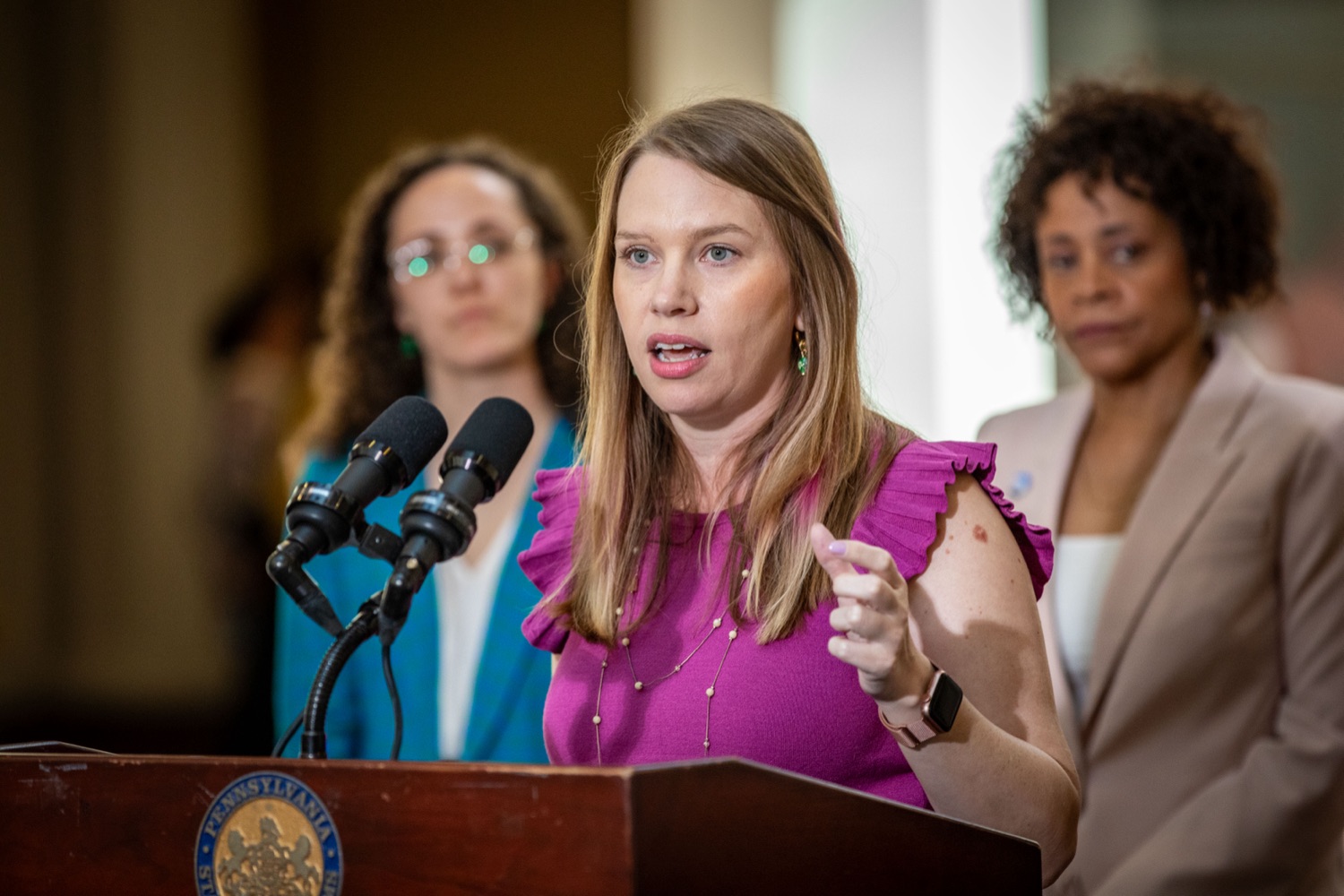 a woman in a pink shirt speaks at a podium, 2 slightly blurry women stand behind her